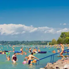 Tłum ludzi cieszy się słońcem i wodą na zatłoczonej Balaton plaży. Dzieci i dorośli pływają, relaksują się na materacach i bawią się w wodzie.