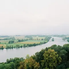 Szeroka rzeka na tle pól i lasów, z mostem w oddali.