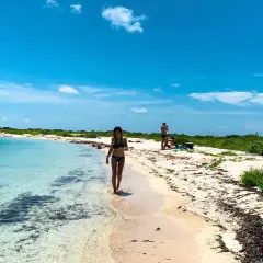 Kobieta spaceruje brzegiem turkusowej wody na plaży w Zatoce Meksykańskiej. W tle widać innych plażowiczów i bujną zieleń.