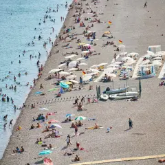 Tłoczna plaża w Finike, ludzie kąpią się w morzu i opalają na kamienistym brzegu pod parasolami.