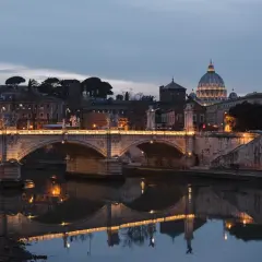 Oświetlony most Ponte Sant'Angelo nad Tybrem, z kopułą Bazyliki św. Piotra w tle. Rzeka w Rzymie odbija światła miasta.