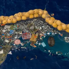 Ogromna wyspa śmieci na oceanie, zebrana w siatkę z żółtymi bojami. Widać plastikowe butelki, sieci rybackie i inne odpady.