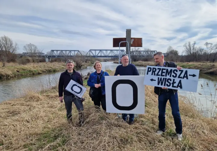 Cztery osoby stoją nad rzeką, trzymając tablice. Jedna z nich głosi "Przemysza Wisła", wskazując na źródło Wisły.