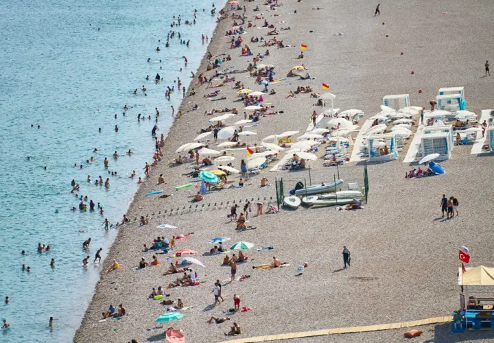 Tłoczna plaża w Finike, ludzie kąpią się w morzu i opalają na kamienistym brzegu pod parasolami.