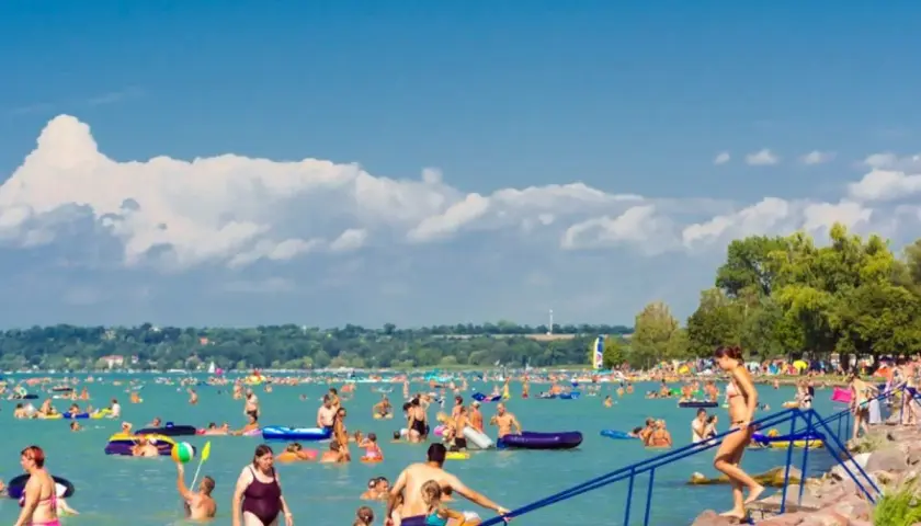 Tłum ludzi cieszy się słońcem i wodą na zatłoczonej Balaton plaży. Dzieci i dorośli pływają, relaksują się na materacach i bawią się w wodzie.