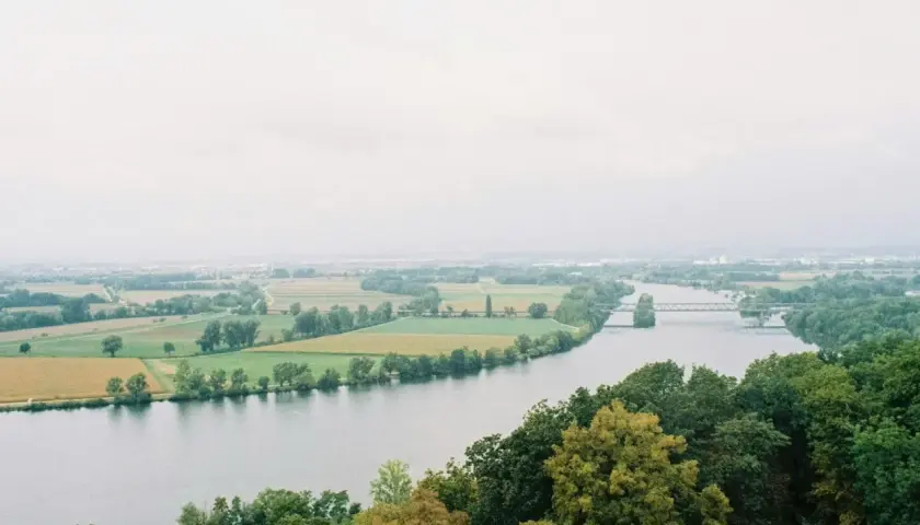 Szeroka rzeka na tle pól i lasów, z mostem w oddali.