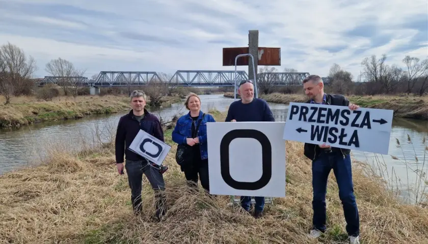 Cztery osoby stoją nad rzeką, trzymając tablice. Jedna z nich głosi "Przemysza Wisła", wskazując na źródło Wisły.