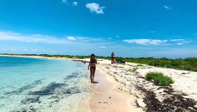Kobieta spaceruje brzegiem turkusowej wody na plaży w Zatoce Meksykańskiej. W tle widać innych plażowiczów i bujną zieleń.