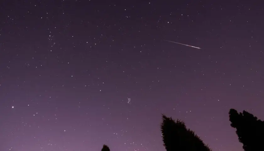 Noc spadających gwiazd. Jasny meteor przecina fioletowe niebo pełne gwiazd, nad ciemnymi sylwetkami drzew.