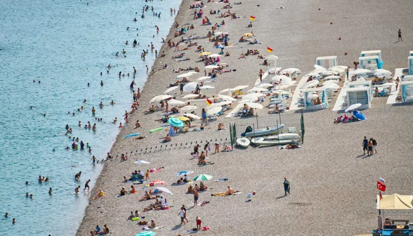 Tłoczna plaża w Finike, ludzie kąpią się w morzu i opalają na kamienistym brzegu pod parasolami.