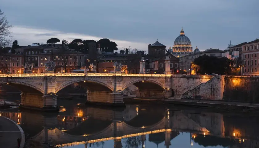Oświetlony most Ponte Sant'Angelo nad Tybrem, z kopułą Bazyliki św. Piotra w tle. Rzeka w Rzymie odbija światła miasta.
