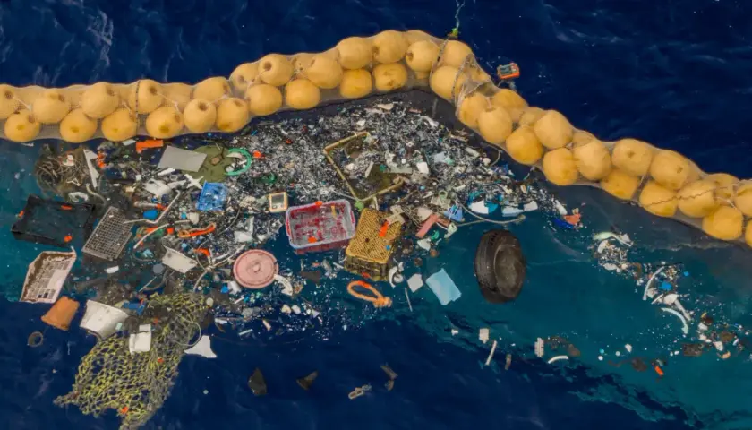Ogromna wyspa śmieci na oceanie, zebrana w siatkę z żółtymi bojami. Widać plastikowe butelki, sieci rybackie i inne odpady.