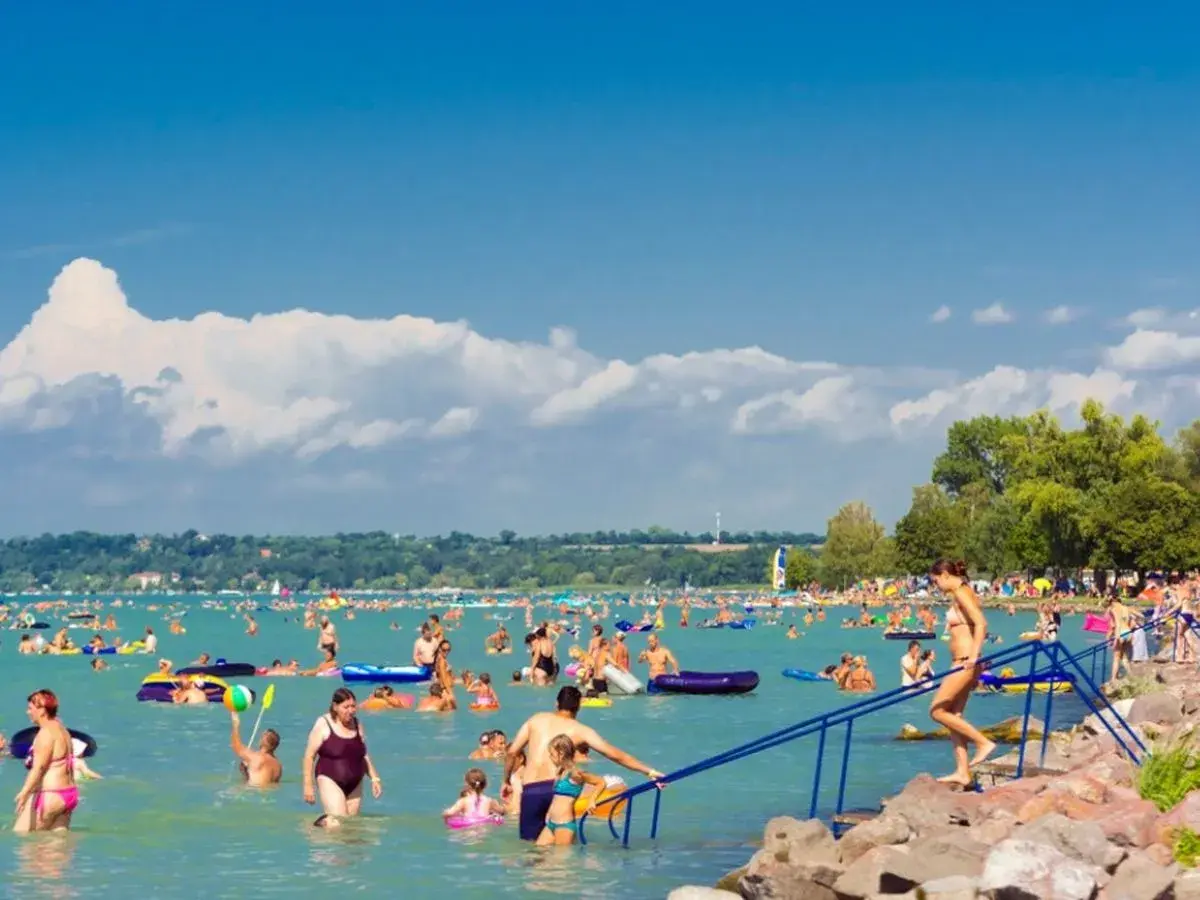 Tłum ludzi cieszy się słońcem i wodą na zatłoczonej Balaton plaży. Dzieci i dorośli pływają, relaksują się na materacach i bawią się w wodzie.