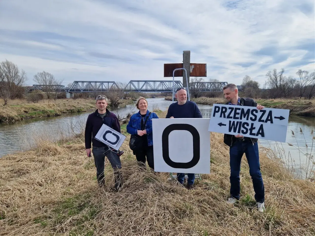 Cztery osoby stoją nad rzeką, trzymając tablice. Jedna z nich głosi "Przemysza Wisła", wskazując na źródło Wisły.