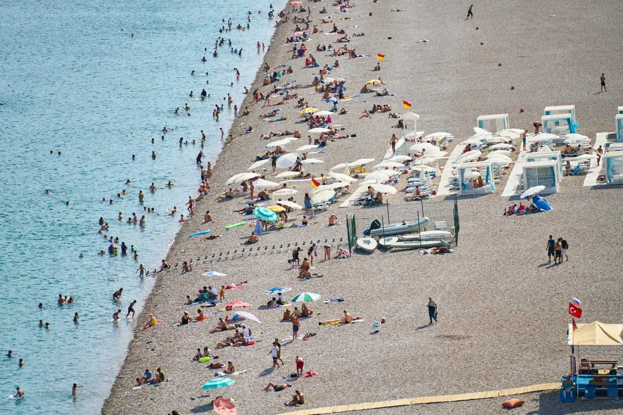 Tłoczna plaża w Finike, ludzie kąpią się w morzu i opalają na kamienistym brzegu pod parasolami.