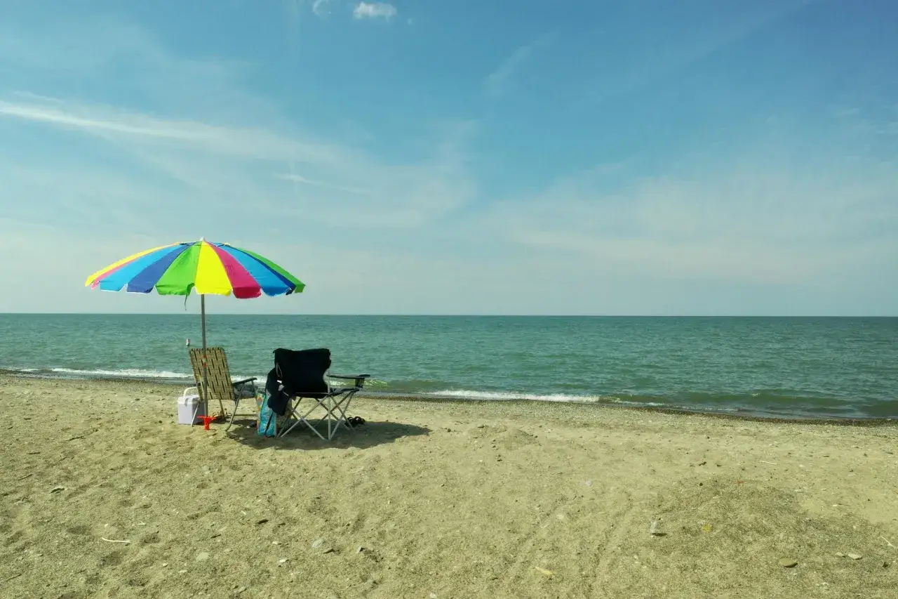 Kolorowy parasol chroni dwa krzesła na piaszczystej plaży nad Balatonem. Woda spokojna, niebo błękitne.