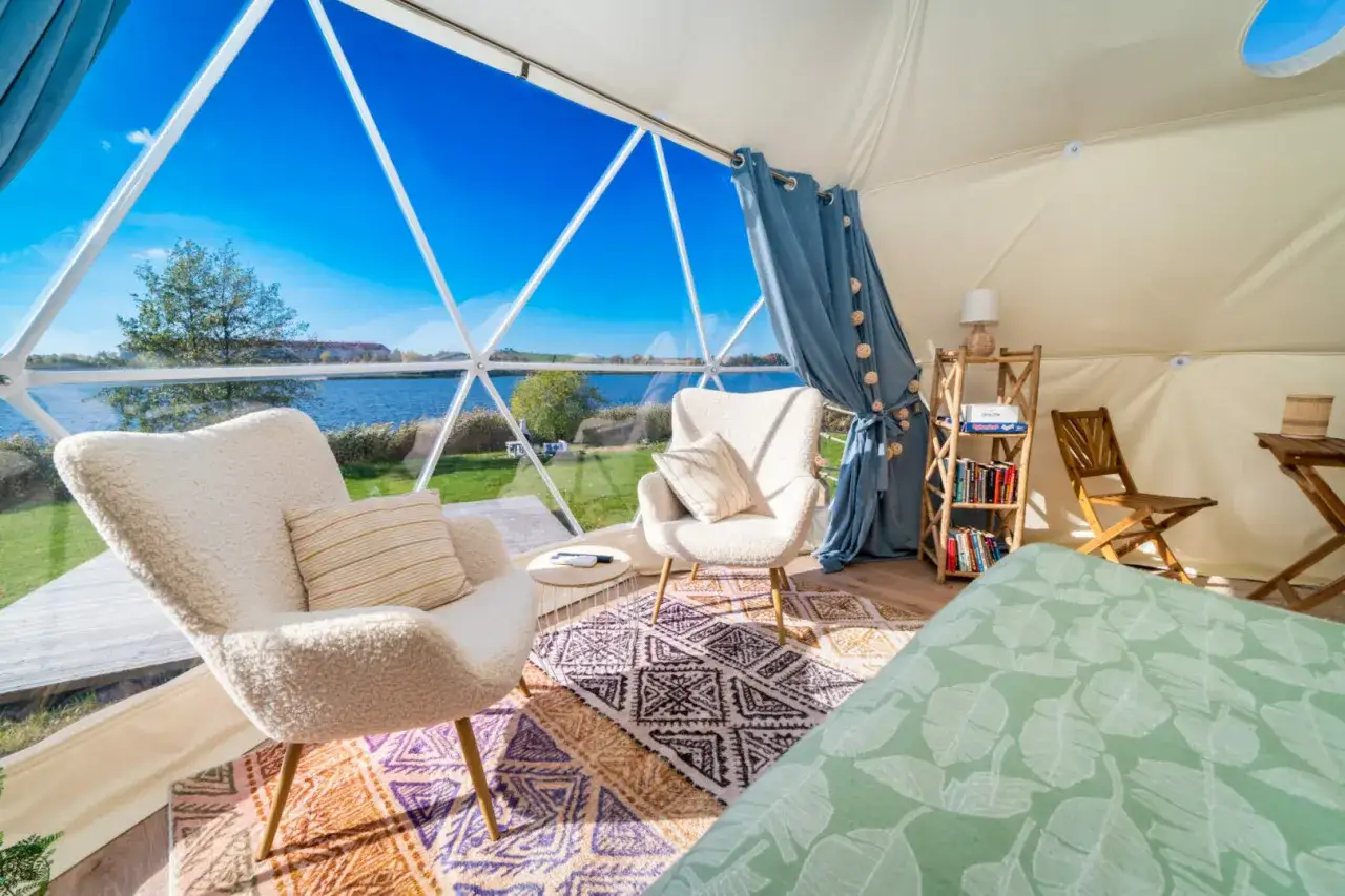 Wygodne fotele i łóżko w namiocie geodezyjnym z widokiem na jezioro. To właśnie glamping co to – luksusowy kemping w otoczeniu natury.