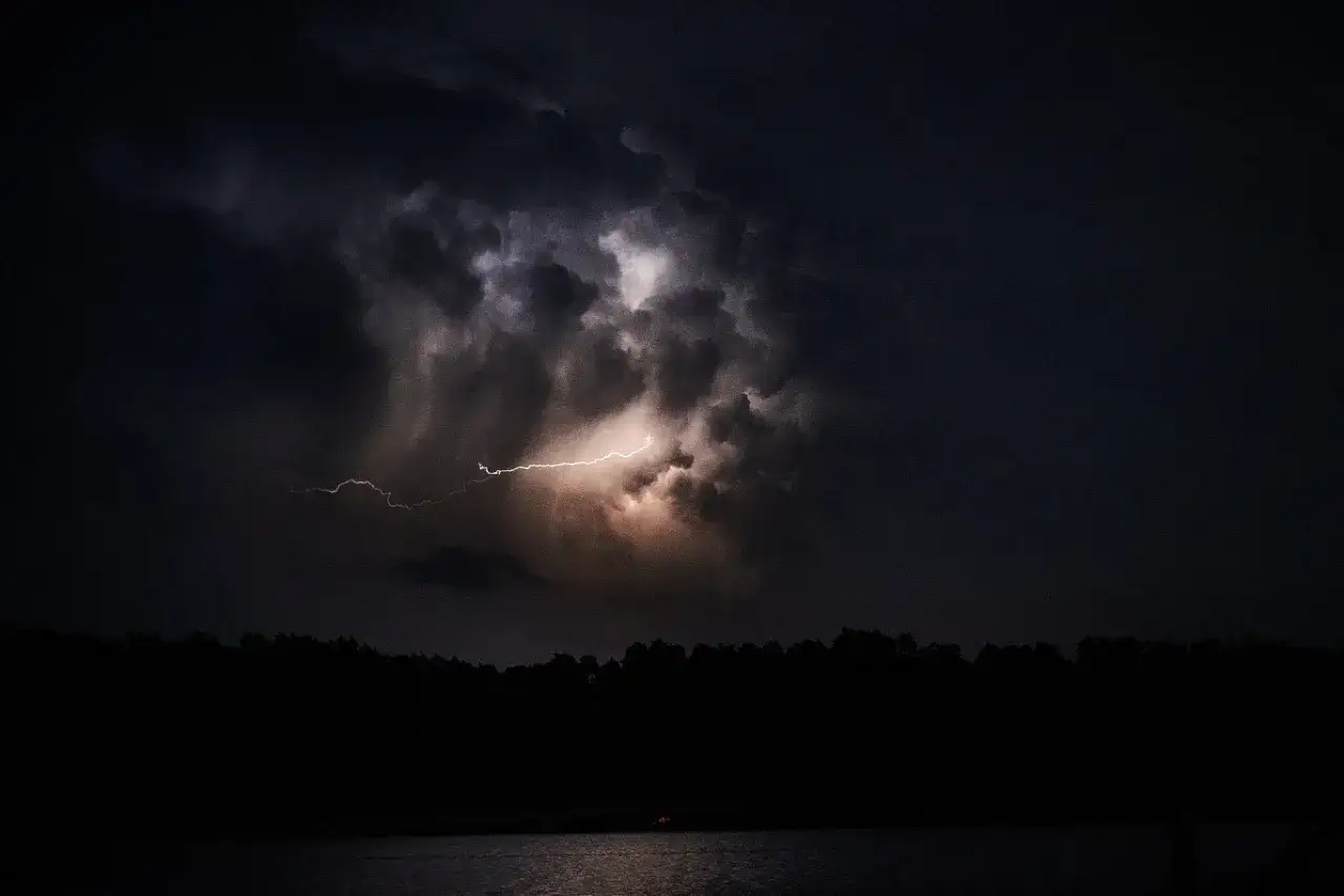 Grzmiąca noc nad jeziorem. Na ciemnym niebie błyska piorun kulisty, rozświetlając burzowe chmury.