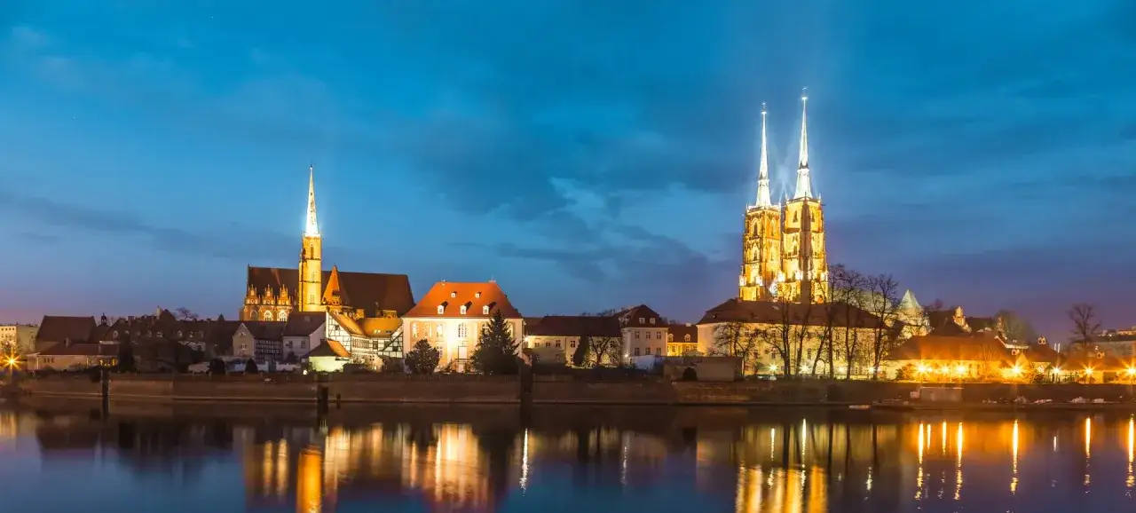 Nocny widok na Ostrów Tumski we Wrocławiu, z oświetlonymi katedrami odbijającymi się w spokojnej Odrze.