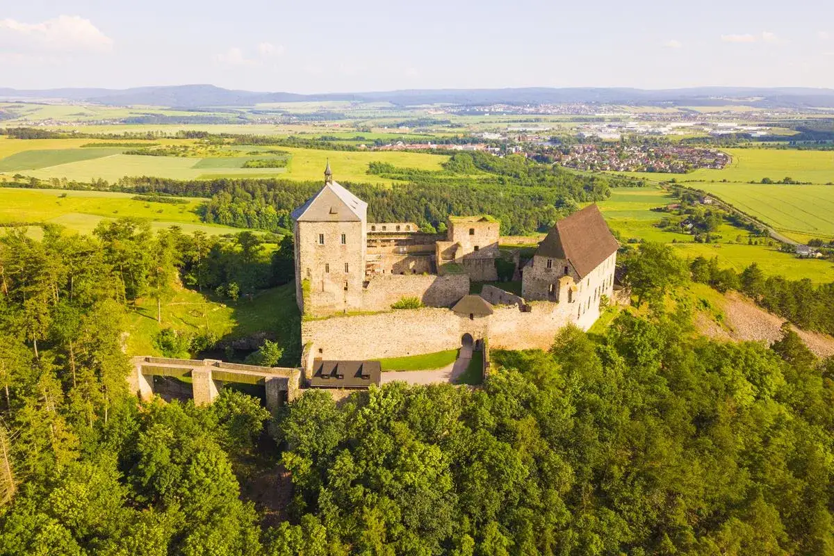 Zamek na wzgórzu, otoczony zielenią, z widokiem na pola i miasteczko. Ciekawe ciekawostki o Czechach często dotyczą takich historycznych miejsc.