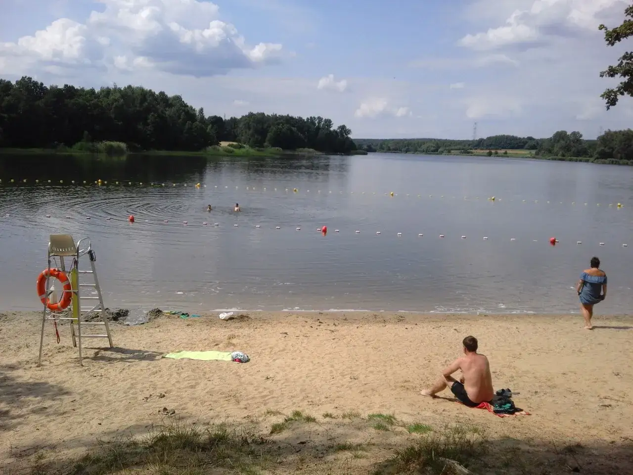 Słoneczny dzień nad jeziorem rybnickim. Na piaszczystej plaży siedzi mężczyzna, a w wodzie kąpie się kilka osób.