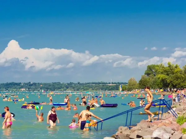 Tłum ludzi cieszy się słońcem i wodą na zatłoczonej Balaton plaży. Dzieci i dorośli pływają, relaksują się na materacach i bawią się w wodzie.